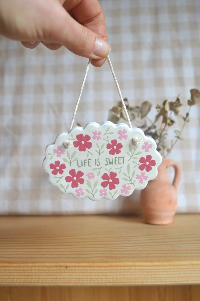 hanging clay sign with red, pink and green floral design reading 'life is sweet' in front of gingham background