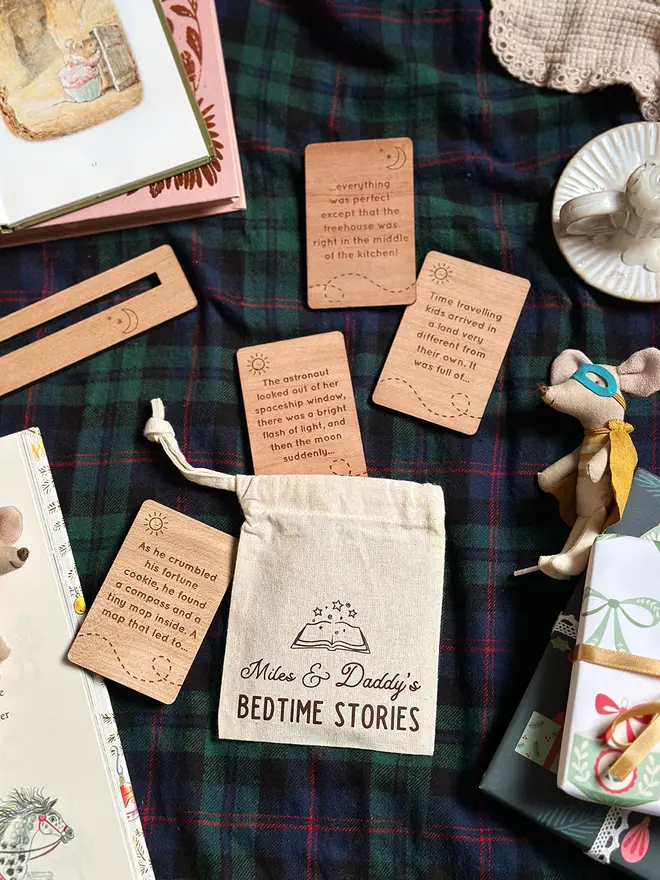 A personalised cotton bag of wooden bedtime story prompt cards is displayed on a cosy red tartan blanket, featuring engraved story ideas. 