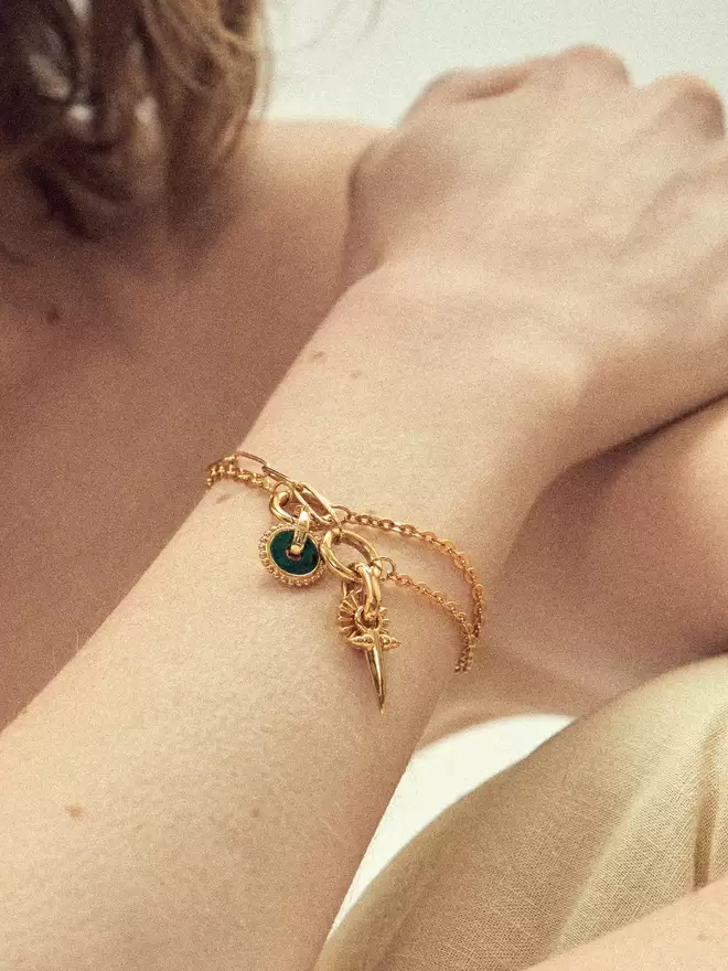 Woman wearing two gold bracelets styled with a malachite disc, a gold Helios disc and a small bayonet charm