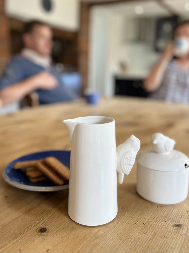 A handmade ceramic sugar pot sits with a jug both with matching bird handles.