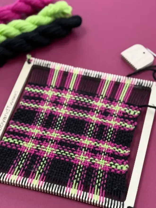 Square frame weaving loom kit with neon pink, green, and black tartan sample, yarn skeins, and wooden tools on a magenta background.