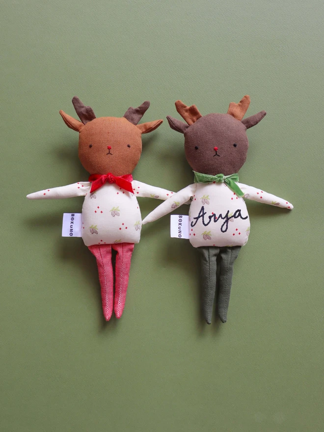 Two handmade reindeer dolls on a green background. Each doll has soft brown antlers, embroidered facial features, and wears a festive white outfit with a pinecone print. One doll has red legs and a red velvet bow; the other has green legs, a green bow, and the name “Arya” embroidered on its body. Both have a "BOKUNO" fabric tag and are positioned holding hands.
