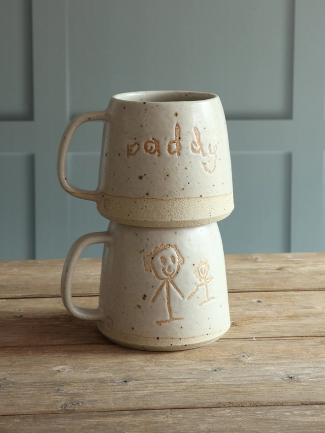 Two Personalised Child's Drawing and Handwriting mugs stacked on top of each other. One mug features a child's stick figure drawing and the second mug has 'Daddy' in a child's handwriting.