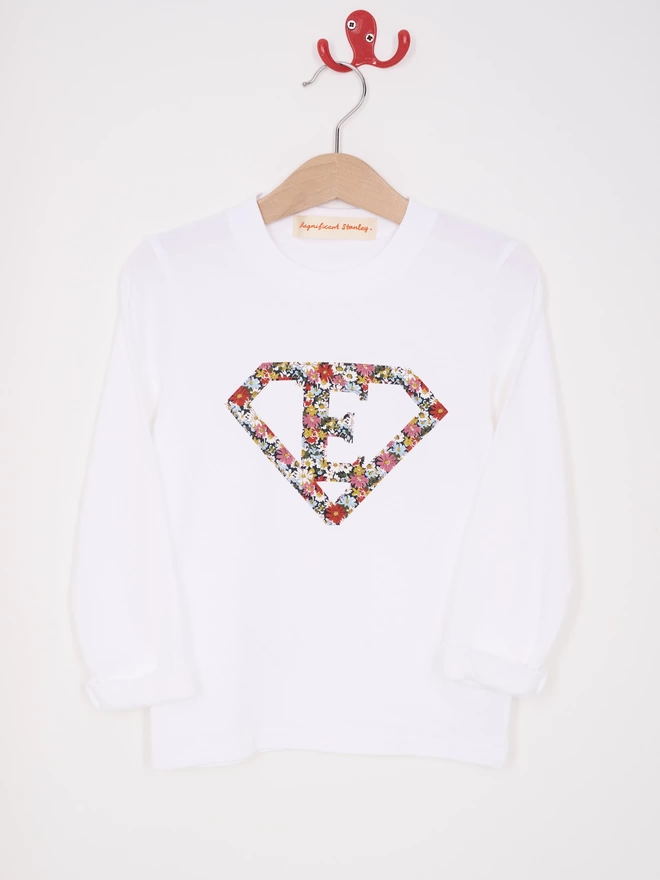 a white long sleeve t-shirt appliquéd with a superhero motif  featuring a letter E initial inside it. The appliqué is made from a floral Liberty print . Hanging on a hanger.