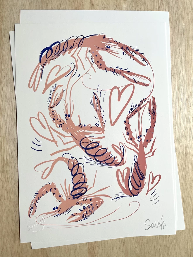 Flat lay of a few sheets of paper showing our pink cartoon langoustine, flung into a frenzied swirl, with hearts and scribbles making this a joyful scene. These are creenprinted in pink with blue accents on a wooden table top.