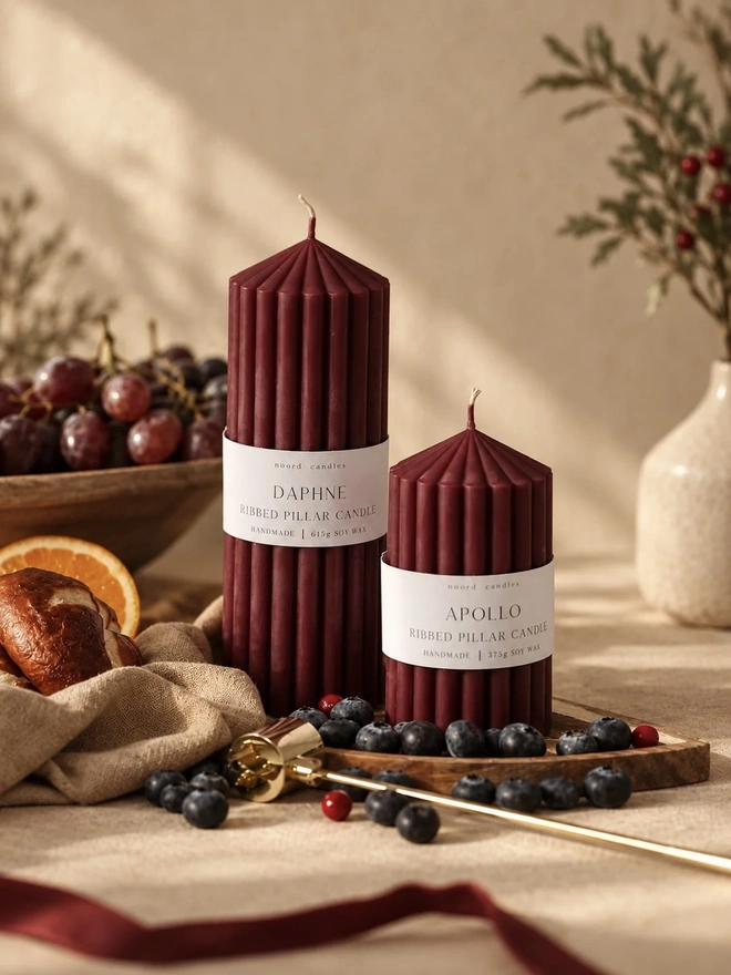 Daphne & Apollo Burgundy Pair Candles (Set Of 2). Two burgundy ribbed pillar candles with pointed tops, wrapped in Noord Candles Daphne and Apollo soy wax labels, styled on a wooden board with blueberries, grapes and a paper honeycomb tree. The rich wine-coloured candles make a stylish centrepiece for festive tables.