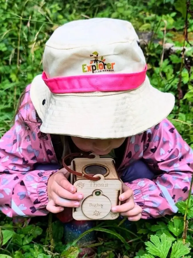 Toddler wearing a white hat and pink raincoat crouches among greenery while observing a Baltic birch plywood bug observation box.
