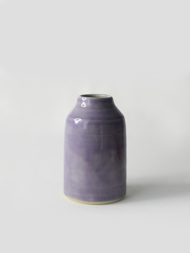 A small handmade ceramic bud vase in glossy amethyst purple glaze, with a rounded bottle shape and narrow neck, photographed against a plain white background