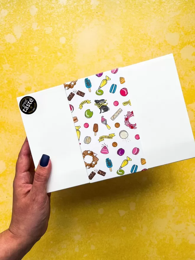 a female hand  with navy blue nails holding up a white gift box with a colourful macaron pattern on it against a bright yellow background