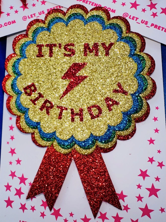 A close up of the 'IT'S MY BIRTHDAY' badge. In the centre, there is a red lightning bolt between the text. The text is on top of layers of scalloped circles in the colours gold, blue, and red. 