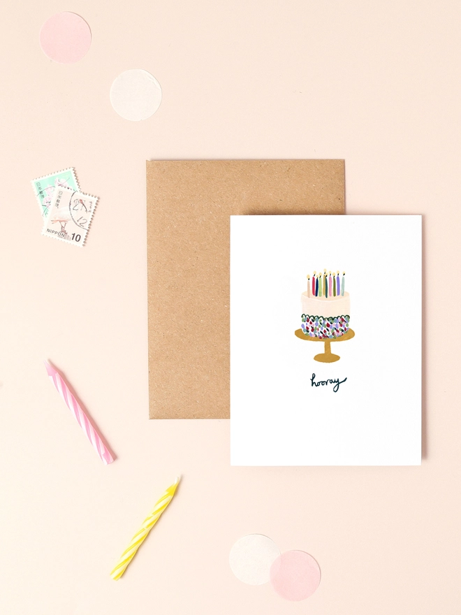 An image of a mini, rectangular white greetings card with an illustration of birthday cake with lot's of candles on a cake stand. A hand written 'hooray' message is seen under the central illustration. The mini greetings card is placed on top of a rectangular brown kraft envelope.