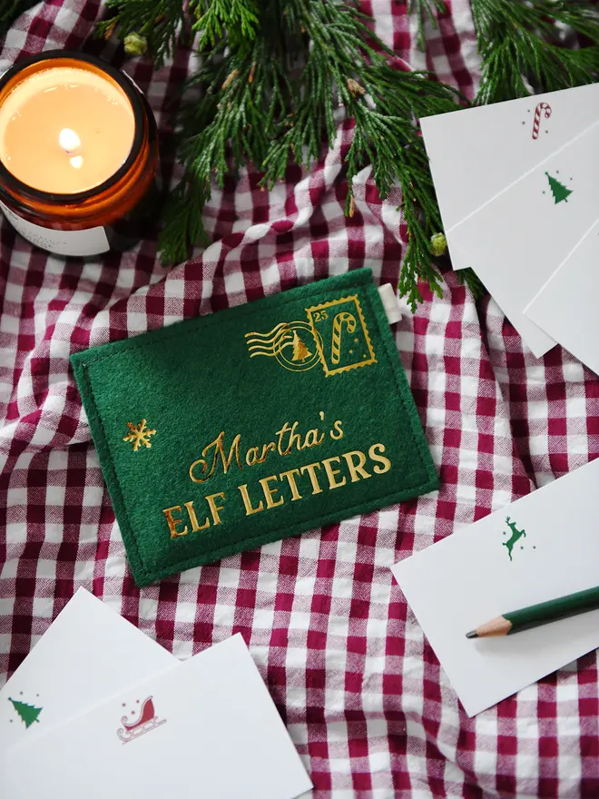 Green felt personalised envelope reading "Martha’s Elf Letters," placed on a red gingham cloth with festive notecards, a green pencil, greenery, and a lit candle.