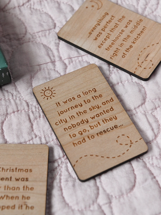Three wooden story cards lay on a pink quilt. Each one has a story idea engraved on it.
