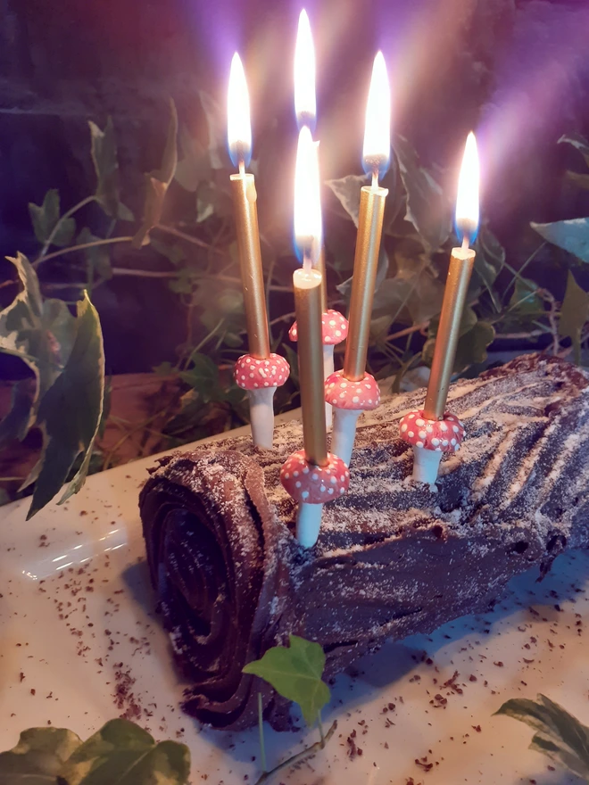 Five handmade ceramic mushroom cake candle holders with red-and-white spotted caps, holding lit gold candles on a chocolate yule log surrounded by ivy.