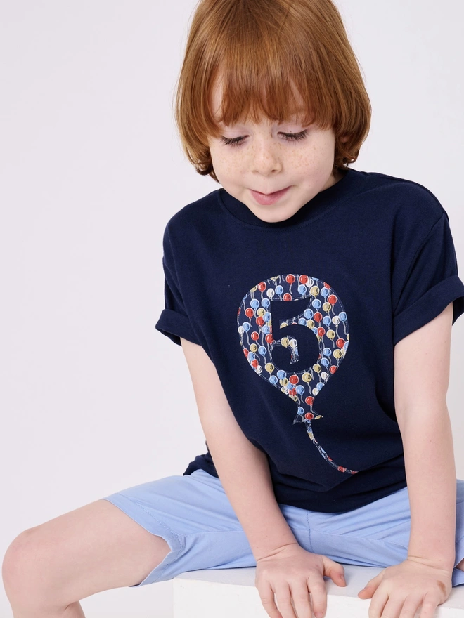 A smiling 5 year old boy wearing a navy cotton long sleeve t-shirt. The t-shirt features a balloon with the number 5 cut out from it, appliquéd in a balloon Liberty print.