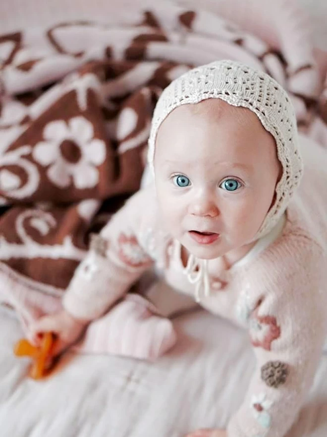 A baby with big blue eyes sits on a bed, staring straight at the camera. The briar rose baby blanket is gathered around her showing the floral pattern.