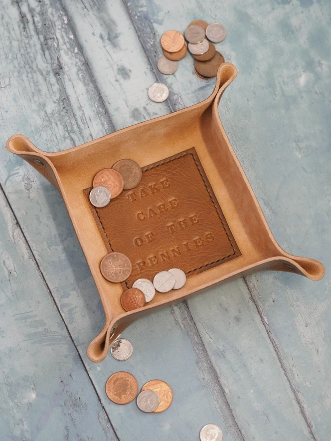 Personalised leather message tray. A square tan brown dish with a square base hand stamped with a personalised message 'take care of the pennies'. Inside the tray are a selection of copper coins and small change.