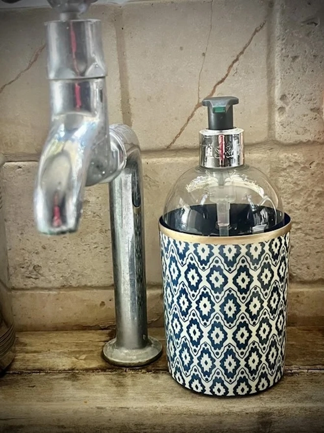 Perfect Enamel Pot. A navy and white geometric patterned pot with a gold rim holding a glass soap dispenser beside a chrome tap. The decorative container adds a stylish, practical touch to a rustic kitchen or bathroom sink.