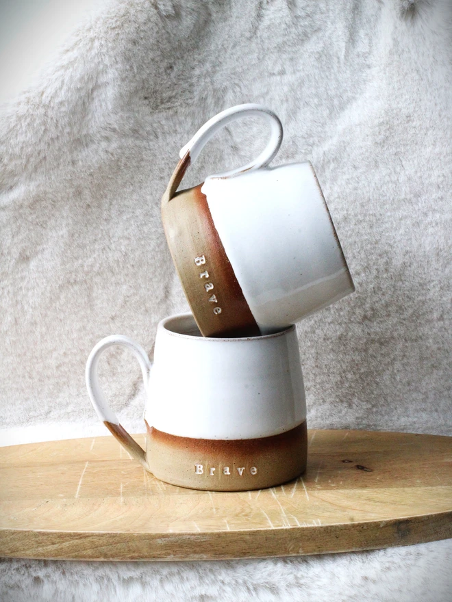 Rustic ceramic mug with a white speckled upper glaze and brown lower half, featuring the word “Brave” on the front, sitting on a wooden table against a neutral background.