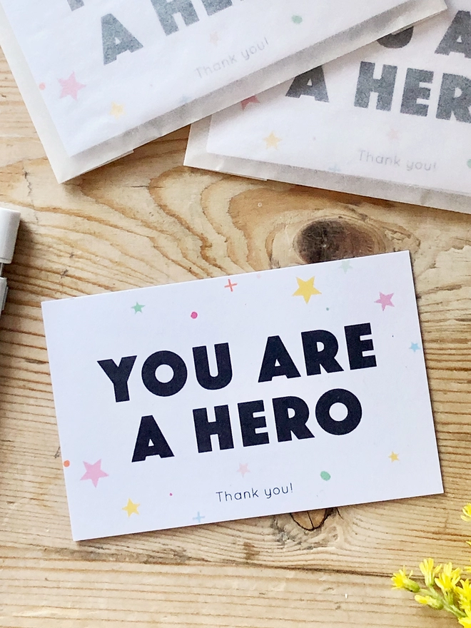 A mini thank you card with the words "You are a hero" and a gentle star design is on a wooden table.