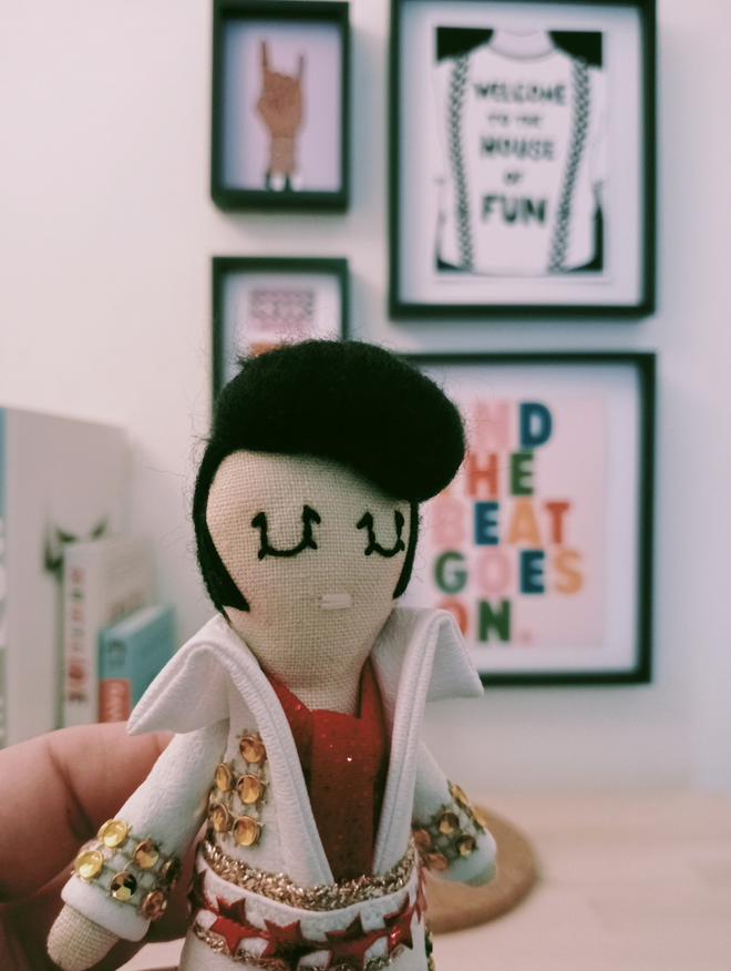 Elvis Presley mini decorative icon doll held up in front of a small gallery wall of music themed prints 