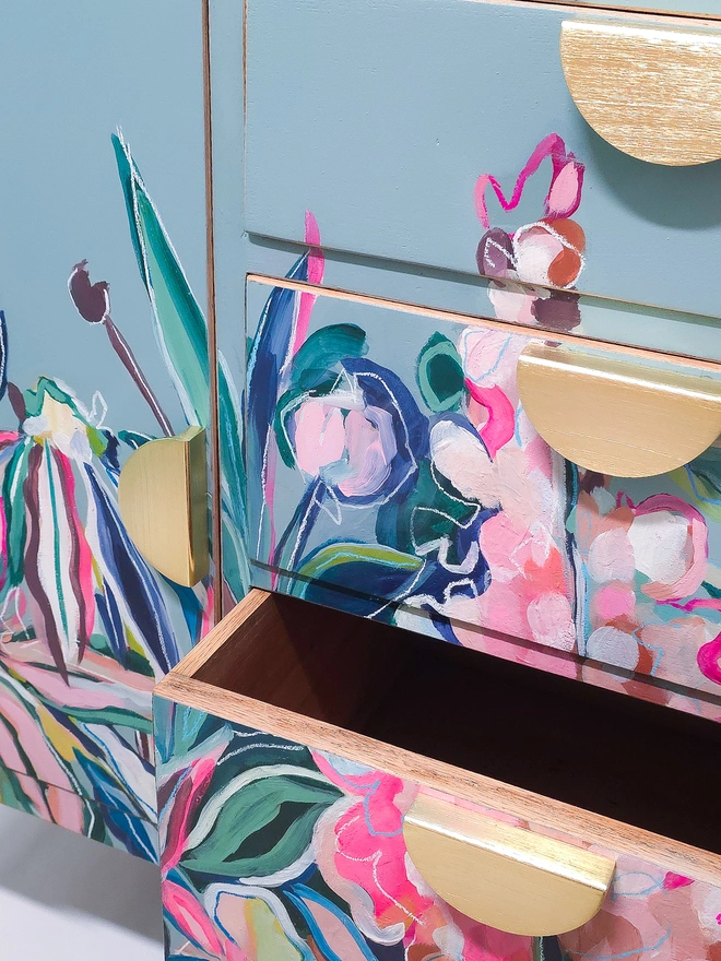 Open Drawer of Vintage Sideboard with brass half-moon handle detail; exterior of sideboard and drawers shows brightly hand-painted floral design in neon pink, green, mustard against a pale dusky blue background
