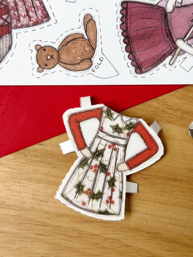 A Christmas card with an illustrated paper doll design is with a red envelope on a wooden desk.