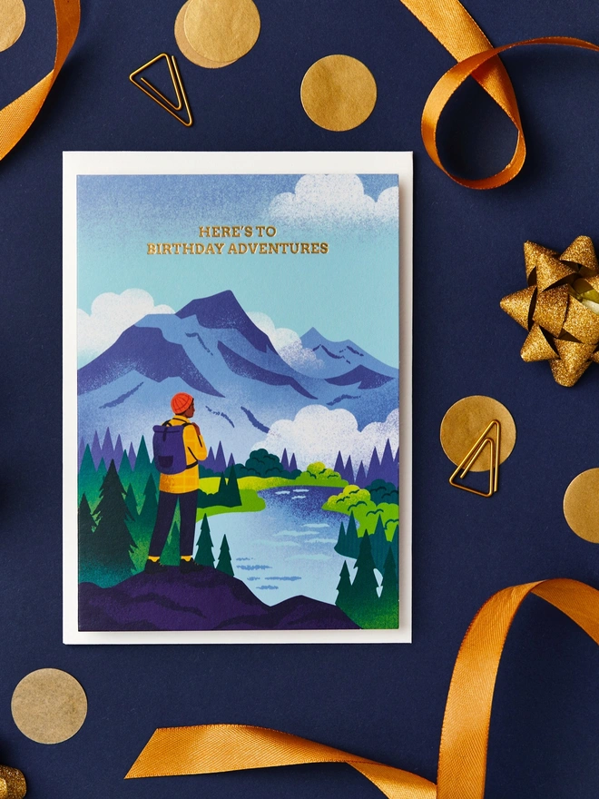 Greeting card with illustration of a hiker on a hill overlooking a lake. The card is on a desk with a navy blue background and gold ribbon decorations.