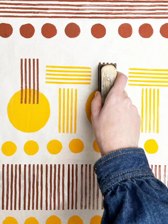 Detail of hand holding a wooden block and printing a yellow stripe onto a yellow and brown geometric design