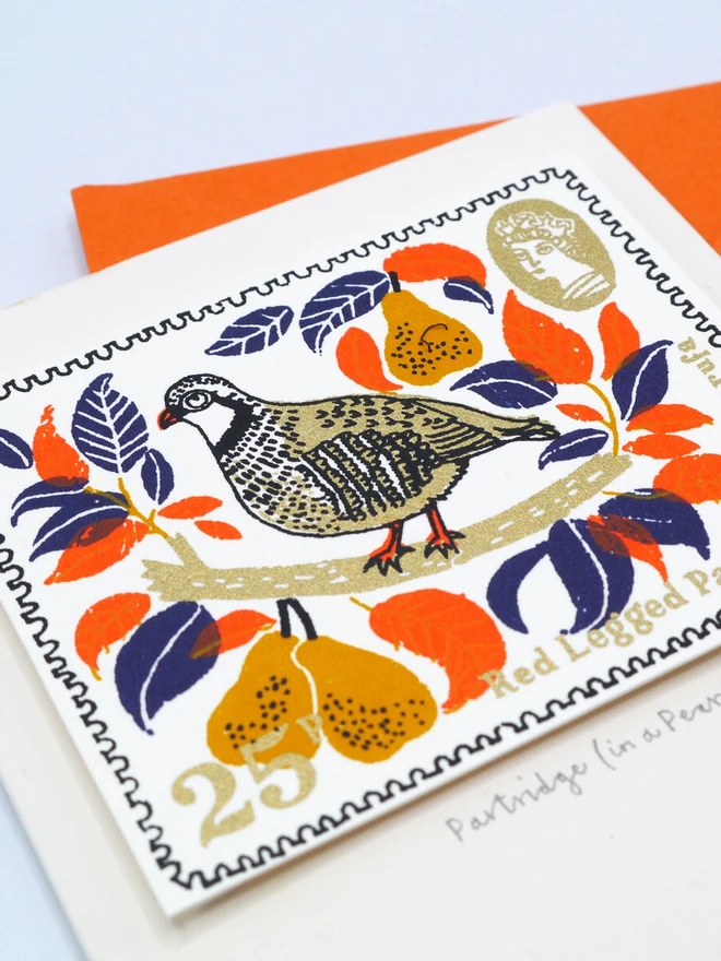 Partridge In A Pear Tree Christmas Card, a white card with a colourful bird design in the centre. It sits on top of an orange envelope on a plain surface. 