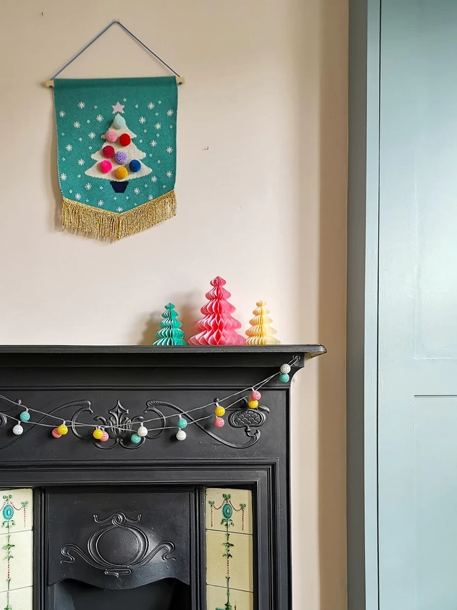 A jade green Christmas tree wall hanging is hung above a Victorian fireplace strung with festive lights and adorned with paper Christams tree decorations on the mantlepiece.