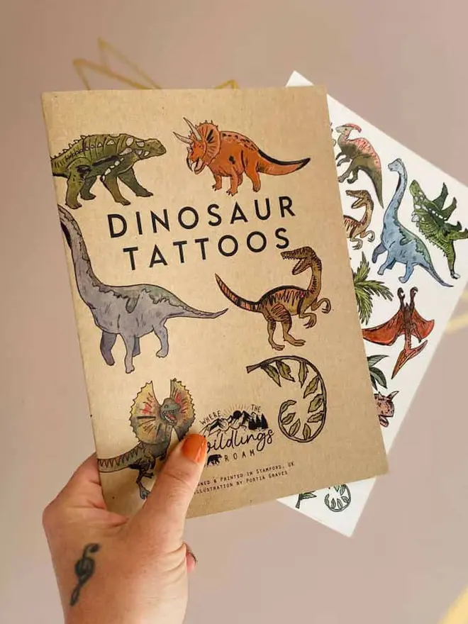 Dinosaur & Fantasy Temporary Tattoos, a hand holds a brown kraft paper booklet titled "DINOSAUR TATTOOS" next to a tattoo sheet with colorful dinosaur illustrations.