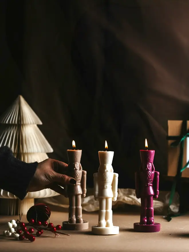 White Nutcracker Candles (Set Of 2). Three lit nutcracker soldier candles in taupe, cream and berry red, with a hand placing one beside a paper honeycomb tree, red berries and a wrapped gift. The traditional