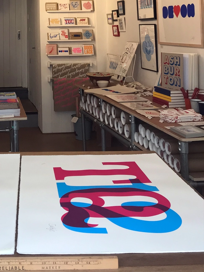 Shop view of a screenprint of TQ12 in overlapping cyan and magenta in the foreground and the shop, shelves, cards and tubes of prints in the background.