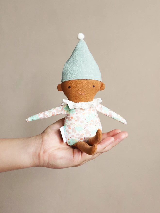 A handmade Spring Elf Doll with brown skin, a multicoloured floral print body in pastel tones, and a pale blue hat with a white pompom. The doll has a cheerful smile and a soft white ruffled collar.