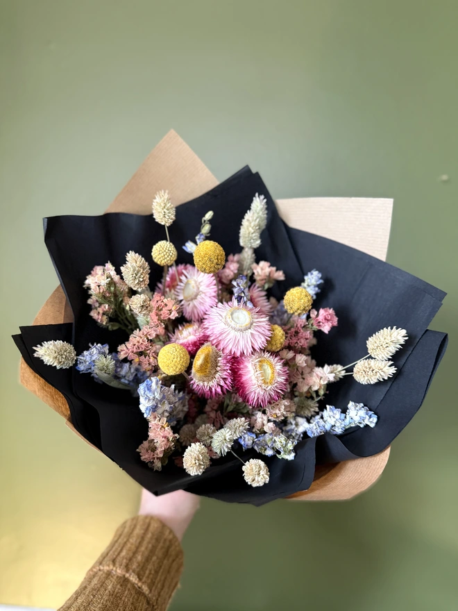 a bouquet of dried flowers in bright colours wrapped in black tissue and kraft paper held by a person in a mustard coloured jumper with their left arm against a green wall