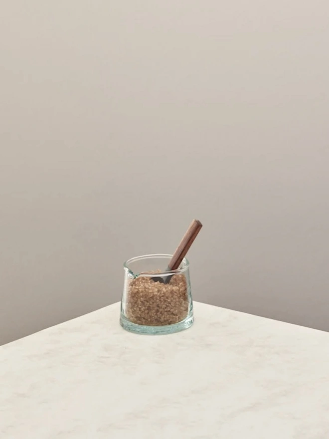 Low Jug, a small glass jug sitting on a counter top against a plain backdrop. It is filled by sugar. 
