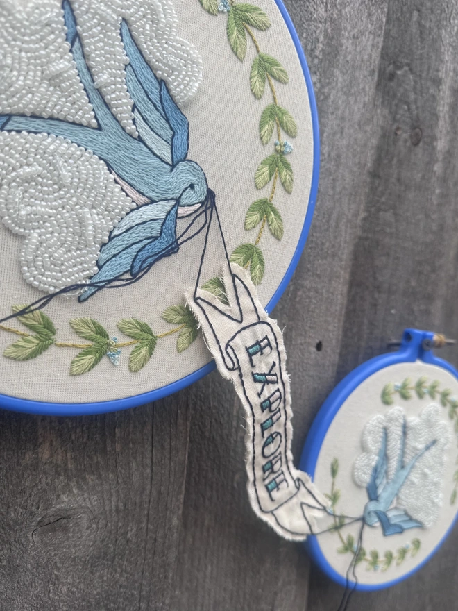 An angled view of two embroidery hoops joined by a banner with the word explore stitched into it held in the beaks of two blue swallows surrounded by y green leaves and in front of white beaded clouds  