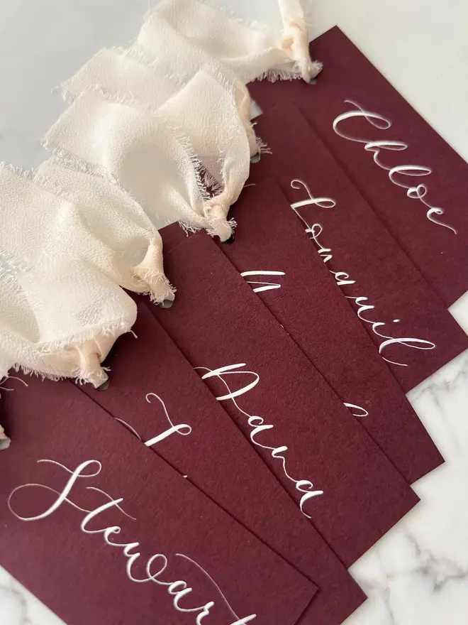 Personalised Calligraphy Wedding Burgundy Place Cards, burgundy place cards set together on a wooden table with champagne ribbons. 