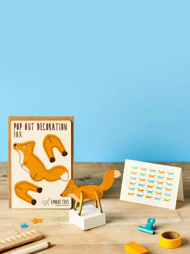 3D laser-cut fox decoration and fox pattern greeting card and brown kraft envelope on top of a wooden desk in front of a sky blue coloured background