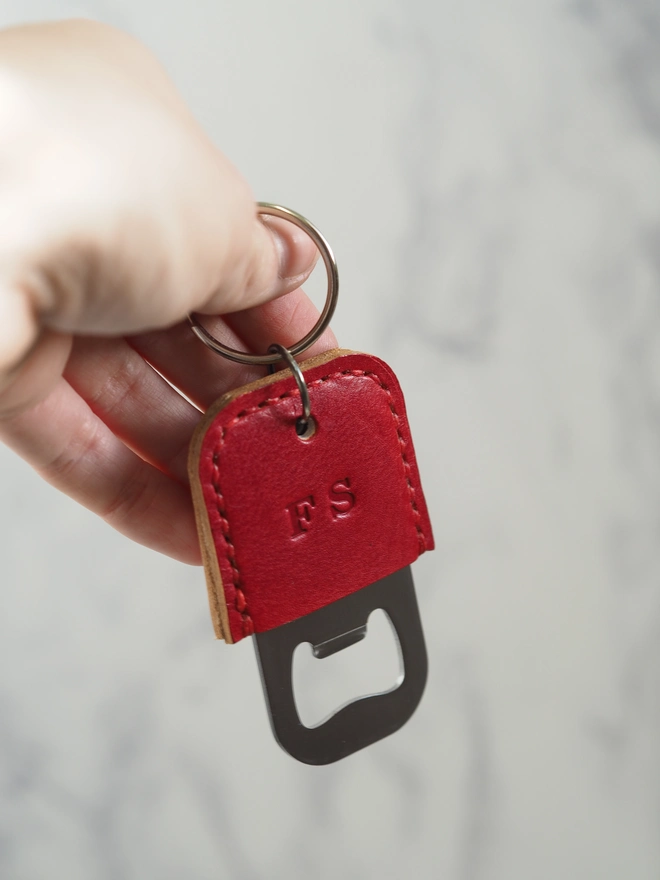 Handmade personalised leather bottle opener key ring. A red bottle opener key chain held in a hand. Personalised with initials in the centre of the leather.