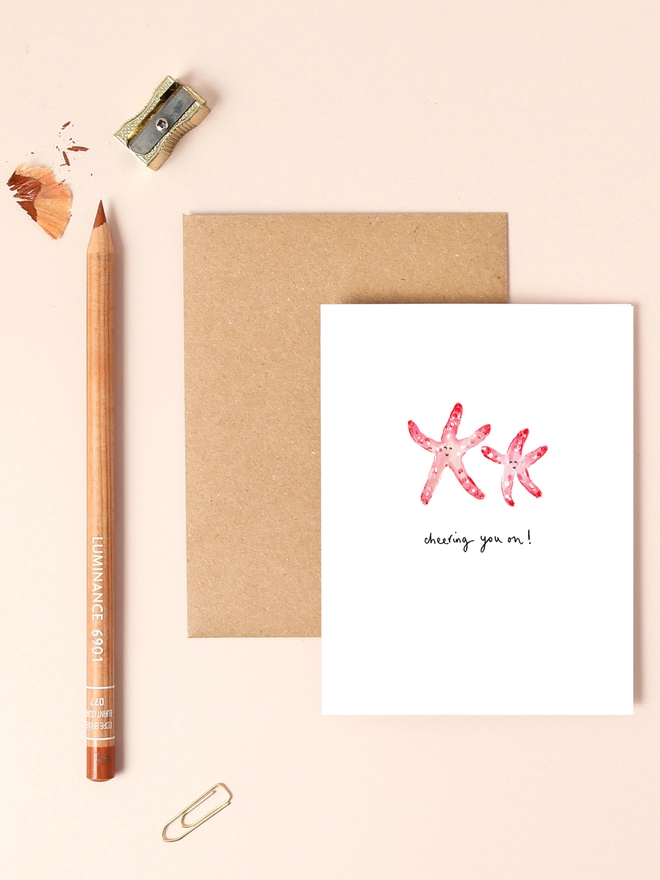 An image of a mini, rectangular white greetings card with an illustration of a couple of pink starfish with smiley faces. A hand written 'cheering you on!' message in black ink seen under the central illustration. The card is placed on top of a brown rectangular kraft envelope.