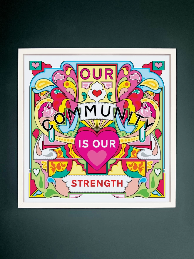 Our Community is Our Strength is written over this bold, symmetrical illustration of yellows, greens and pinks. The print is in a white square frame hung on a dark grey wall. 