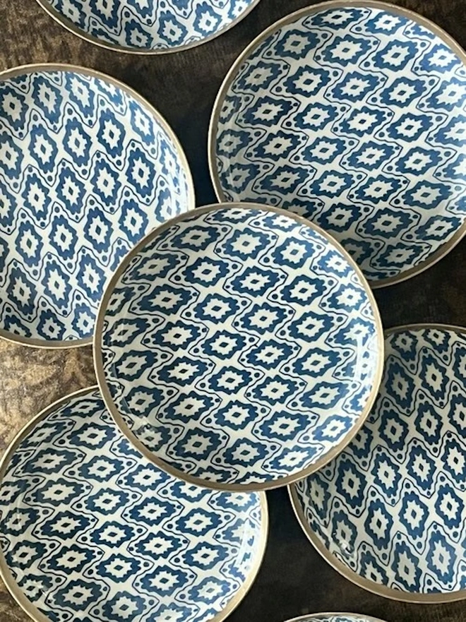 Small Enamel Circular Trinket Tray. A collection of navy and cream ikat-patterned dishes with gold rims arranged on a dark textured surface. The matching geometric designs make a striking set for decorative storage or gifting.