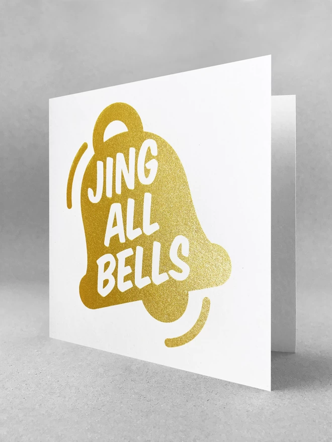 Slightly open, a sparkly gold ink graphic of a bell with the words Jing All Bells within. A square card in a studio set. Handprinted in ashburton 