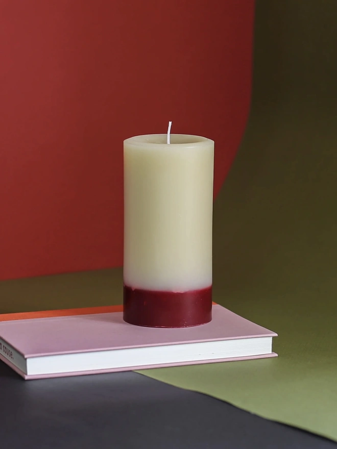 Oudh, Yuzu & Patchouli Candle (Set Of 1 Or 3 Wicks). the oudh, yuzu and patchouli candle sits on a pink book against a red and olive green background, showing its single wick and cream wax with a deep red base. the two-tone colouring and textured, slightly uneven wax surface give the candle a rustic, hand-poured finish. the bold, contrasting background colours highlight the candle's warm tones.