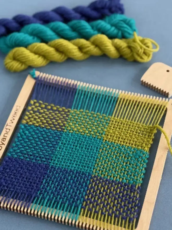 Square wooden frame weaving loom kit with blue, teal, and yellow woven sample, matching yarn skeins on a blue background.