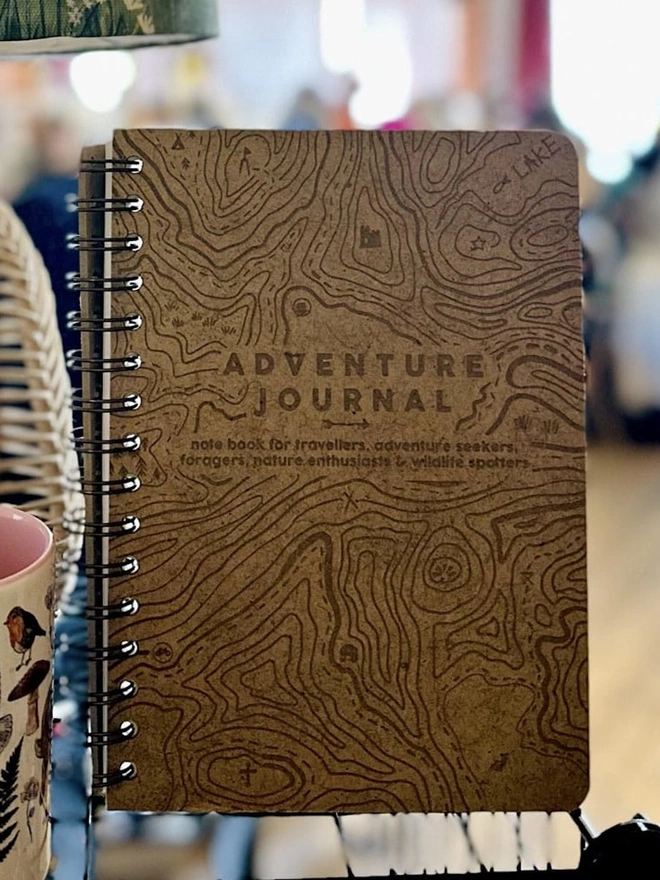 Wooden Hardback Adventure Journal, an adventure journal sitting in a shop stand accompanied by other products. 