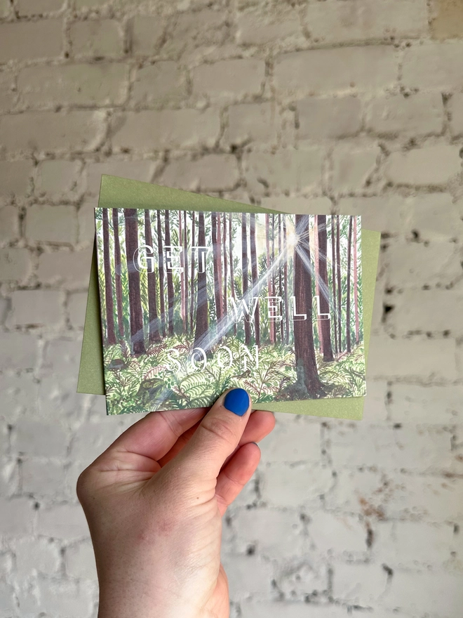 a greetings card featuring an illustration of a woods landscape with the text “get well soon” with a green envelope