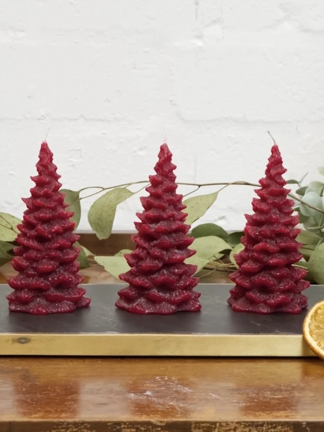 Green Or Red Christmas Tree Candle (Set Of 3). three red christmas tree shaped candles stand in a row on a slate and brass tray, set on a wooden surface with eucalyptus foliage and dried orange slices arranged around them. each candle has a textured, layered pine branch design tapering to a point, with a wick at the top. the rich red wax and festive styling, with its natural greenery and citrus, give the set a warm, seasonal feel suited to holiday decorating.